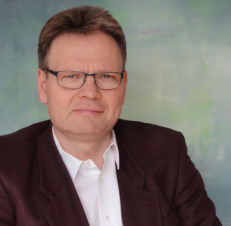 Ein Mann sitzt am Tisch und bereitet Unterlagen für ein Gespräch vor. Im Hintergrund verlaufen grüne und blaue Farbbahnen.